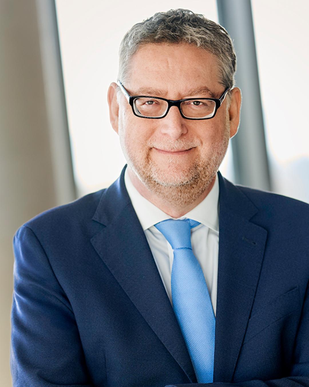 Porträt eines Mannes mit kurzer grauer Frisur, schwarzer Brille und Bartstoppeln im dunkelblauen Anzug, weißem Hemd und hellblauer Krawatte vor hellem, unscharfem Hintergrund.
