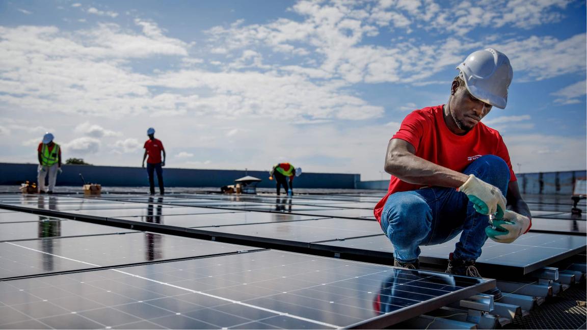 Ein Arbeiter in Helm und Handschuhen montiert Solarmodule auf einer großen Dachfläche, während weitere Personen im Hintergrund arbeiten.