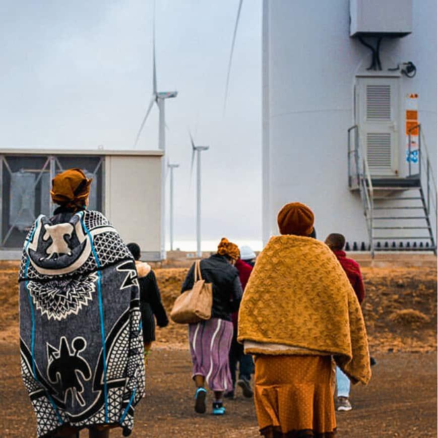 Eine Gruppe von Menschen in warmen Umhängen geht auf eine Windkraftanlage zu, während weitere Windräder in der Ferne zu sehen sind.