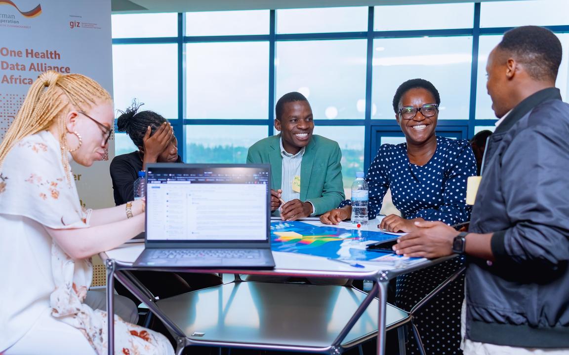 Mehrere Personen sitzen in einem hellen Raum an einem Tisch mit Laptop und Unterlagen und sprechen miteinander, im Hintergrund ein Banner „One Health Data Alliance Africa“.
