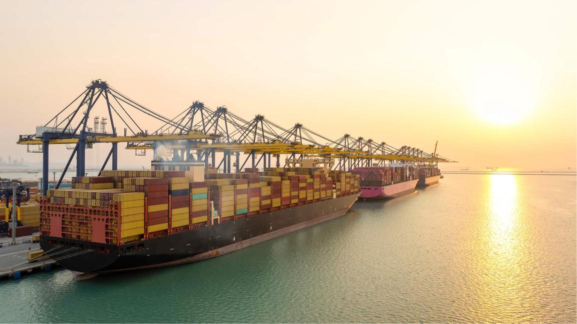 Ein Containerschiff liegt bei Sonnenuntergang an einem Terminal und wird von mehreren Hafenkränen beladen, während Container in Reihen gestapelt sind.