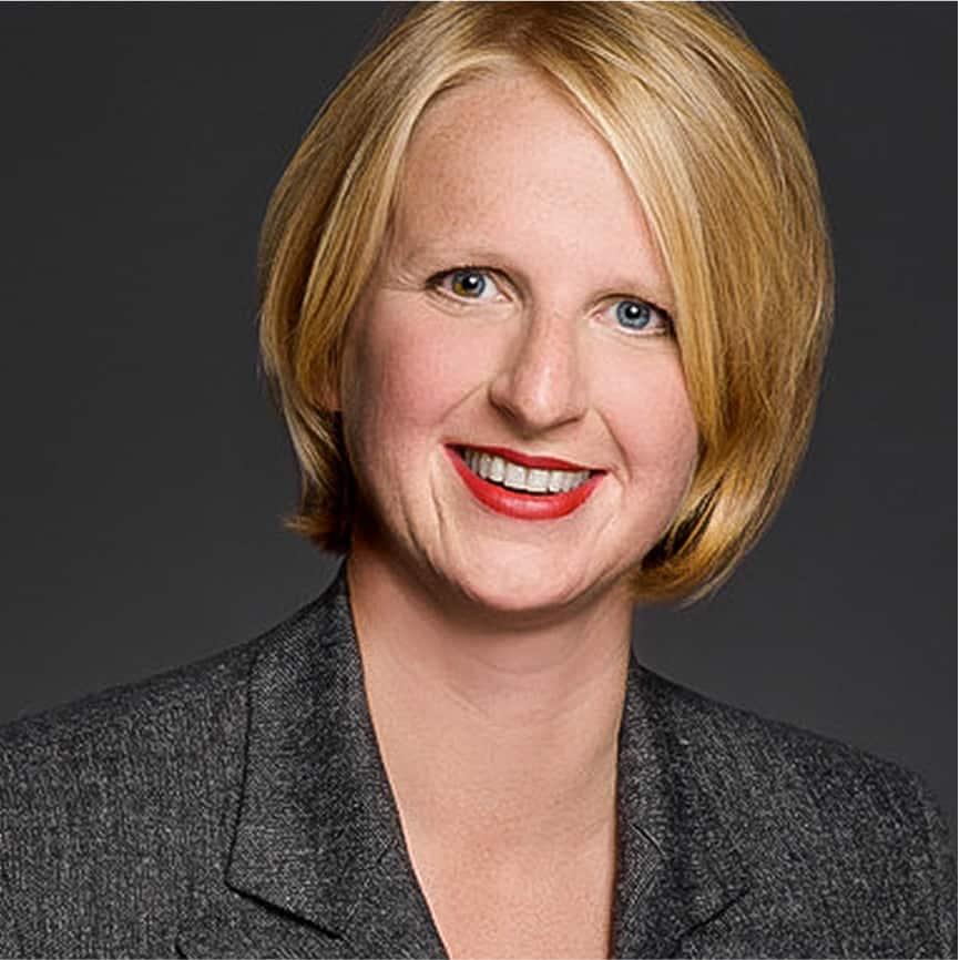 Porträt einer lächelnden Frau mit blondem Bob, rotem Lippenstift und grauem Blazer vor dunklem Hintergrund.