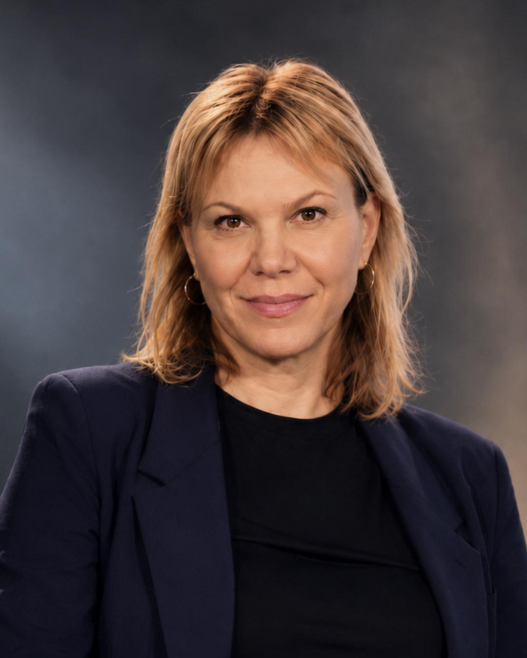 Studio-Porträt einer Frau mit schulterlangem blondem Haar, dunklem Oberteil und dunkelblauem Blazer vor dunklem Hintergrund.
