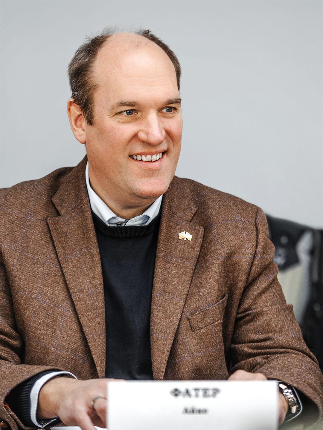 Portrait of a smiling man in a brown blazer against a light background, seated at a table.
