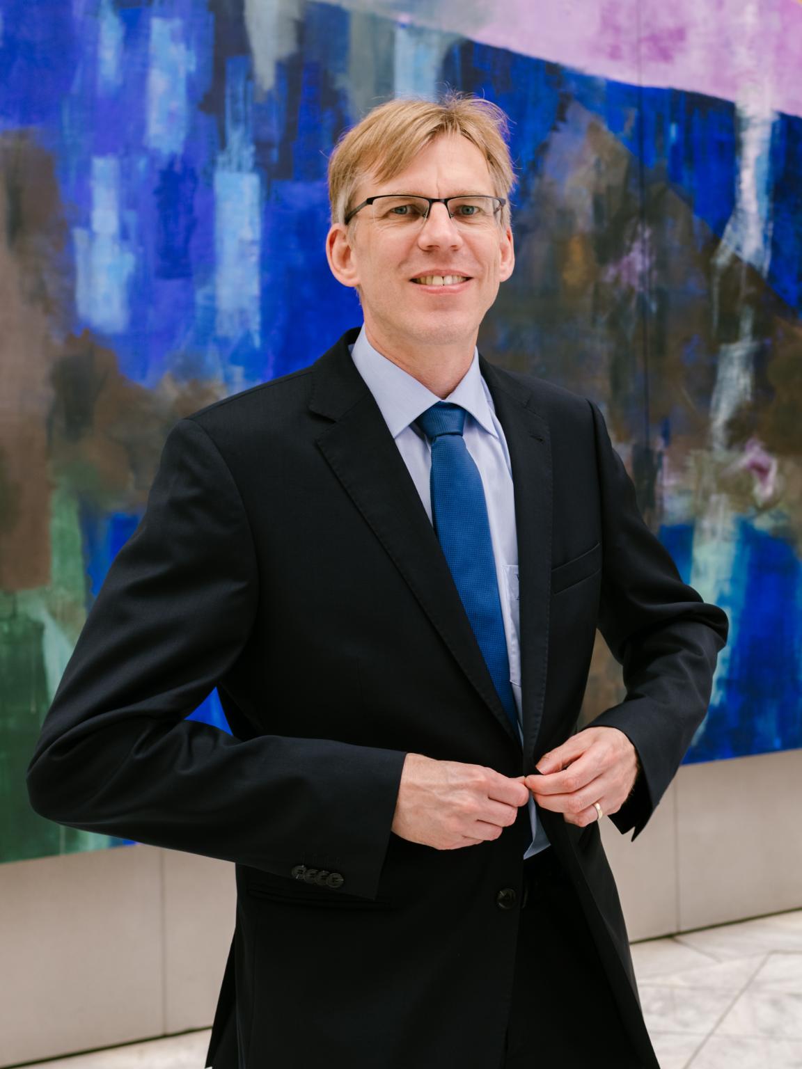 Portrait of a man in a dark suit with a blue tie and glasses in front of a large abstract blue-purple painting.