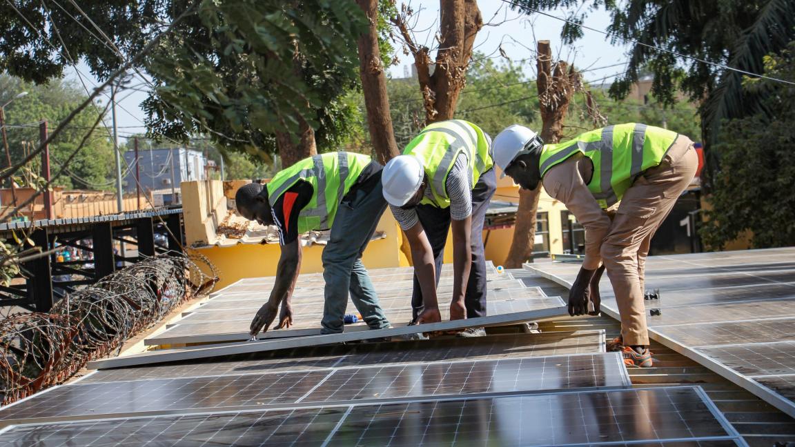Three people in high-visibility vests and hard hats install solar panels on a roof.