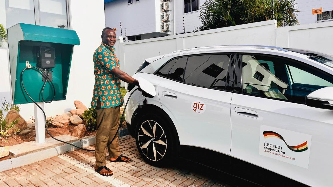 A person charges a GIZ electric car at a charging station in a parking space in front of a building.