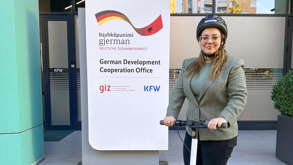 A person on an e-scooter in front of a sign for the German Development Cooperation Office with the GIZ and KfW logos.