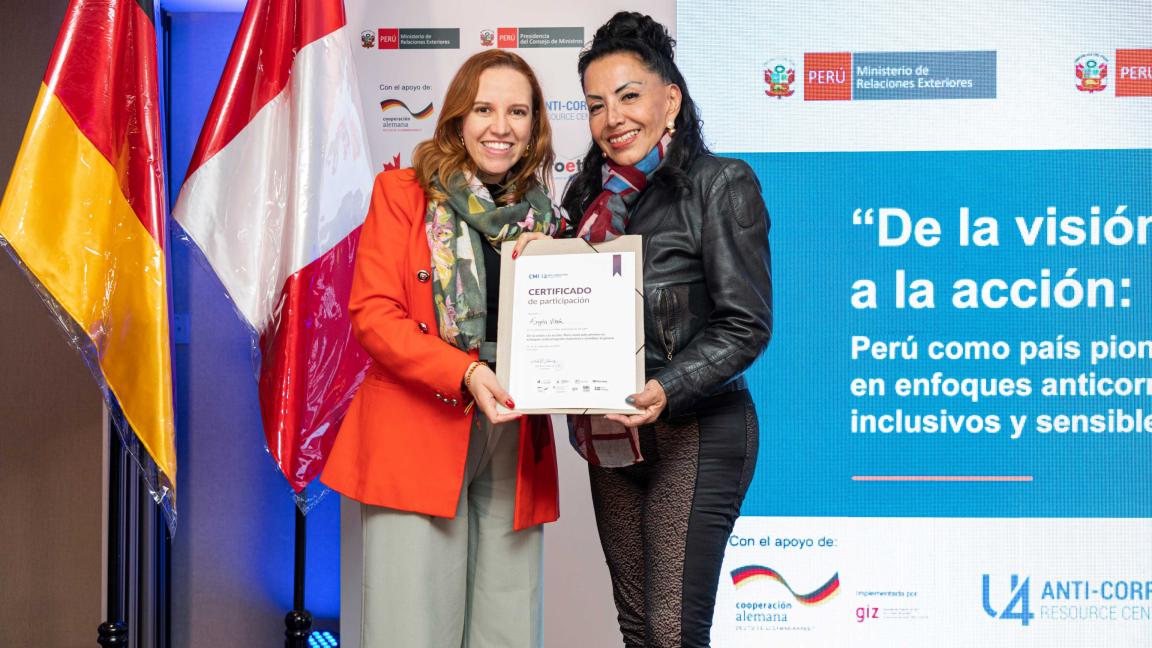 Two people stand on a stage with the German and Peruvian flags and hold a certificate.