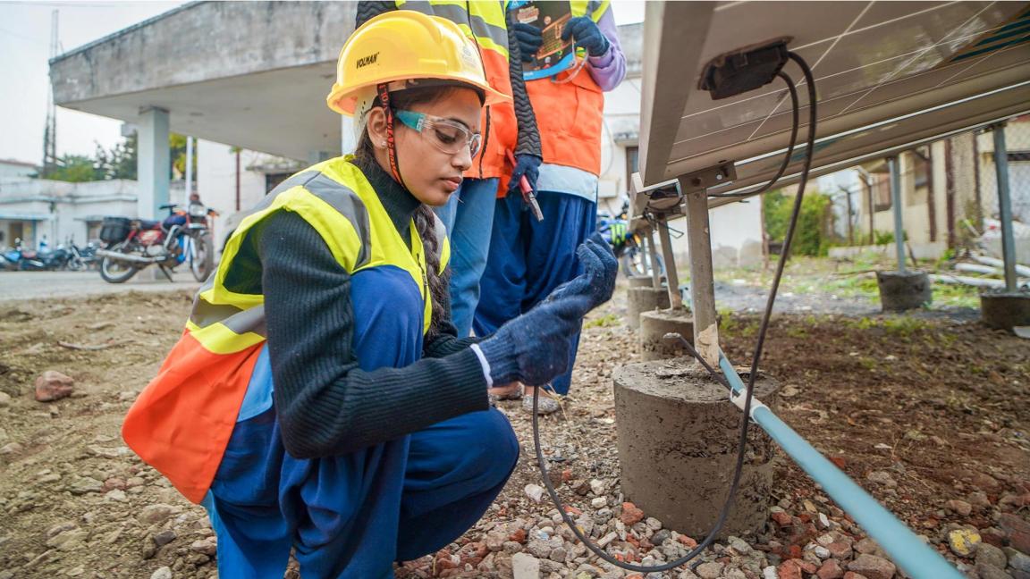 A young woman wearing protective clothing checks the cables under a solar panel; alongside her are two other people with tools and documents.