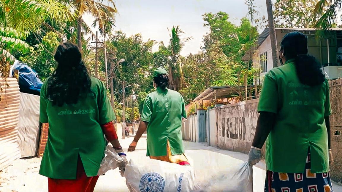 Three women in work clothes walk along a street carrying a full refuse bag.
