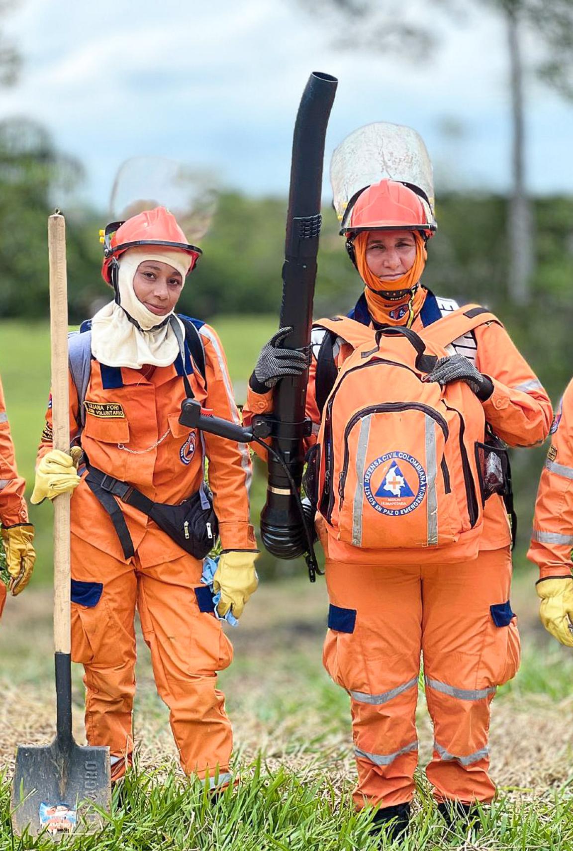 Two workers in orange protective clothing and helmets stand in a meadow holding shovels and a motorised blower. 