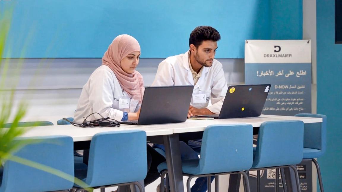 Zwei Personen arbeiten konzentriert an Laptops in einem modernen Raum mit blauen Farbakzenten. Beide Personen tragen weiße Arbeitskittel.