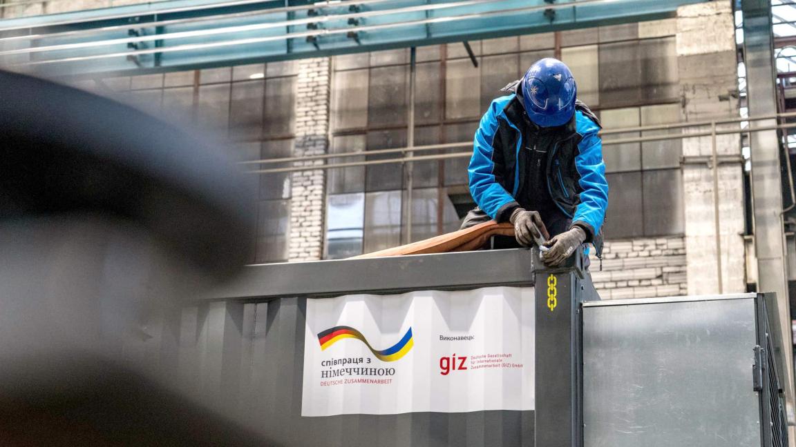 Eine Person mit Schutzhelm arbeitet auf einem Container, auf dem ein Banner mit GIZ-Logo und deutsch-ukrainischer Zusammenarbeit hängt.