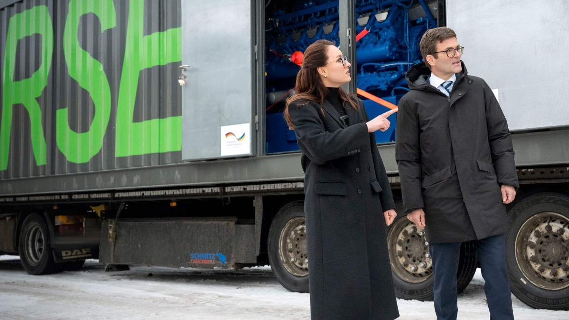 Eine Frau und ein Mann stehen vor einem Lkw mit Container und geöffneter Technikklappe, durch die eine blaue Maschine sichtbar ist.
