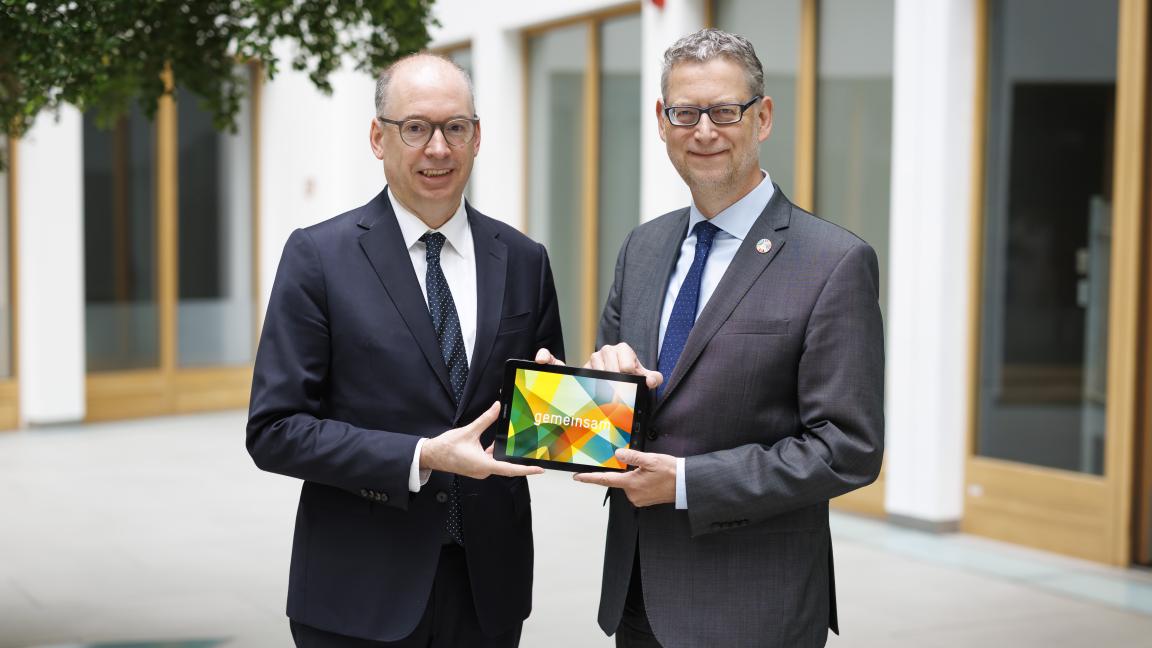 Zwei Männer in Anzügen und mit Krawatte halten gemeinsam ein Tablet mit bunter Grafik und dem Schriftzug Gemeinsam in den Händen