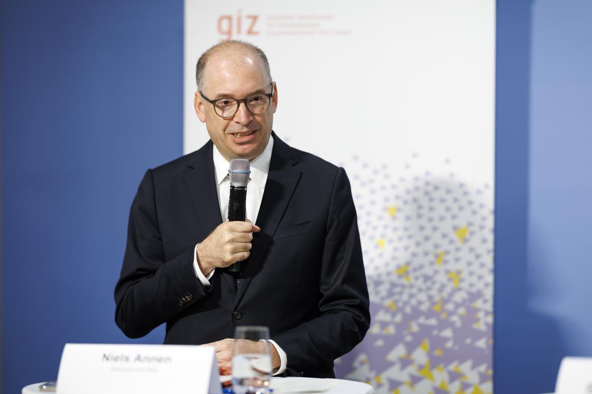 A man in business attire is speaking into a microphone in front of a roll-up banner bearing the GIZ logo. In front of him is a name plate bearing the name Niels Annen