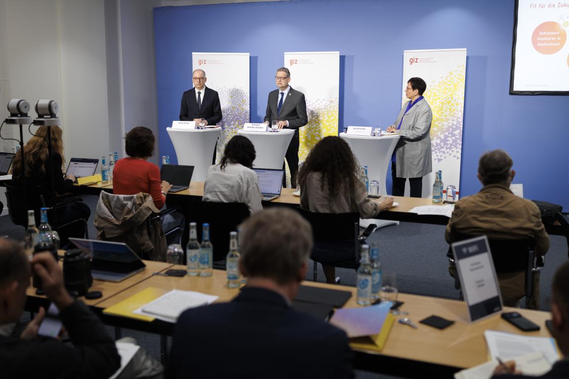 Ein Raum mit zwei Tischreihen, an denen sieben Menschen sitzen. Sie blicken auf eine Bühne, auf der drei Menschen an Rednerpulten stehen.