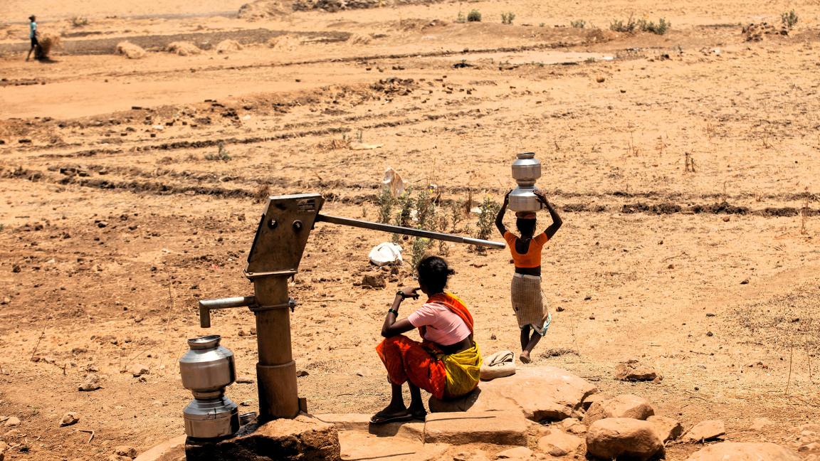 Zwei Frauen an einer Handwasserpumpe in einer trockenen, sandigen Landschaft, eine sitzt neben Metallwasserbehältern und die andere trägt einen gestapelten Behälter auf dem Kopf.