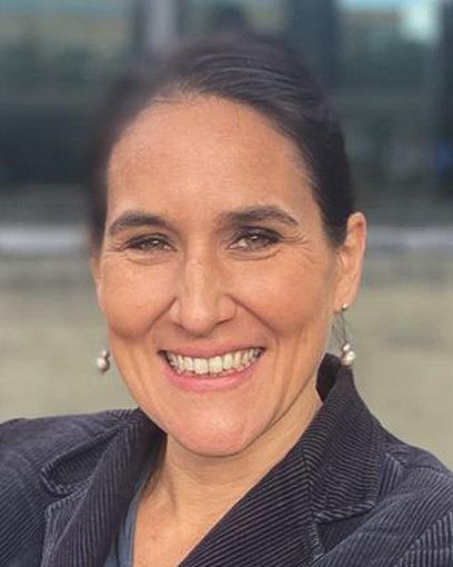 Portrait of a smiling woman with hair tied back, wearing a dark jacket, outdoors against a blurred background.