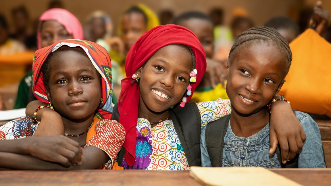 Three schoolgirls sit side by side in a classroom, smiling at the camera, wearing colorful headscarves and school backpacks.