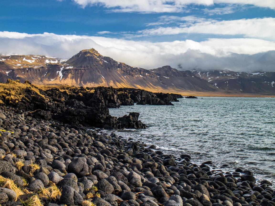 Steiniger Küstenabschnitt mit dunklen Lavasteinen am Ufer, ruhigem Wasser und schneebedeckten Bergen unter tiefhängenden Wolken im Hintergrund.