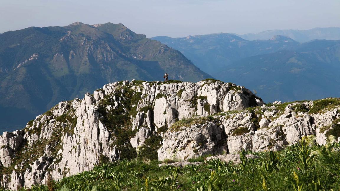 Eine Person wandert auf einem hellen Felsgrat, während dahinter blau dunstige Bergketten in die Ferne reichen.