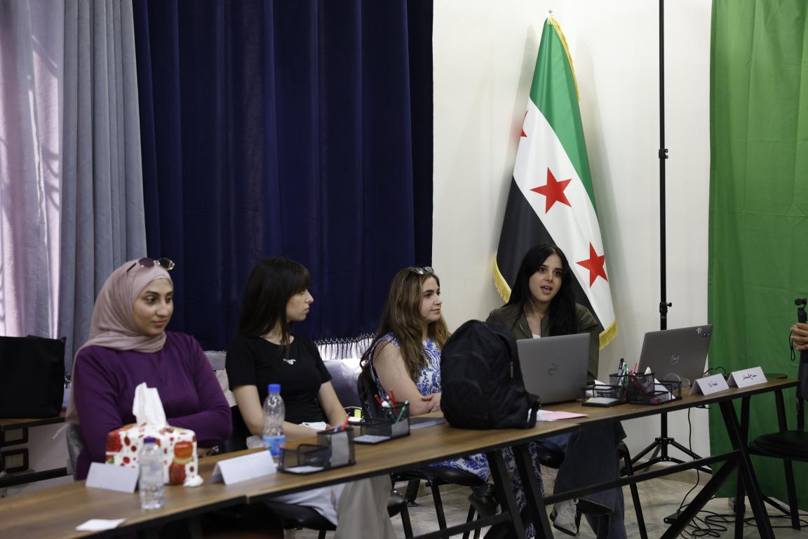 Vier der Teilnehmerinnen sitzen an einem Tisch vor einer syrischen Flagge