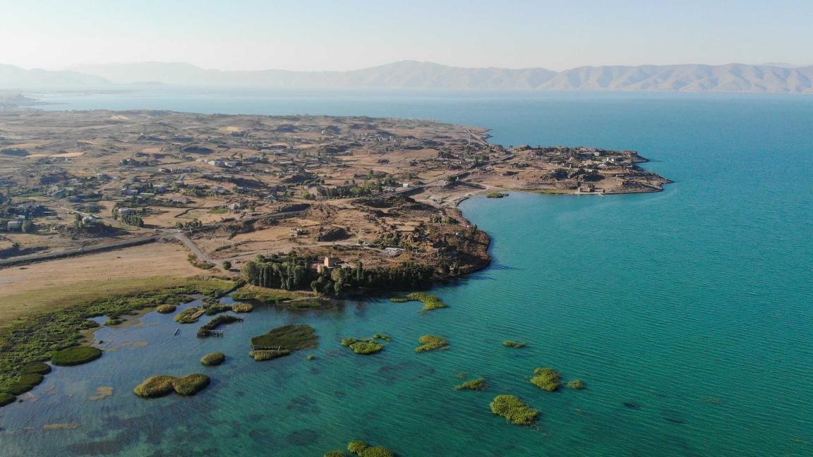 Luftaufnahme einer kargen Küstenlandschaft mit Bucht, türkisblauem Wasser und kleinen grünen Inselchen im Vordergrund sowie Bergen am Horizont.
