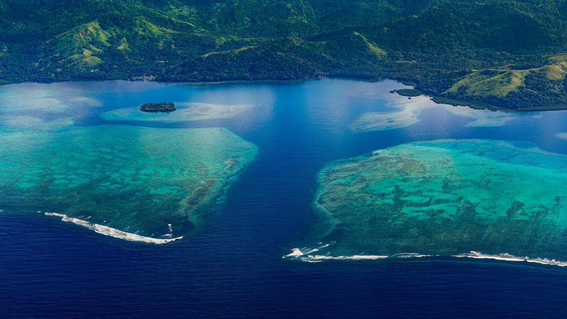 Aerial view of a coastline with two turquoise reefs separated by a deep blue channel, with forested green hills in the background.