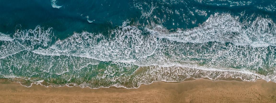 Aerial view of ocean waves breaking with white foam onto a golden-brown sandy beach, with turquoise to dark blue water in the background.