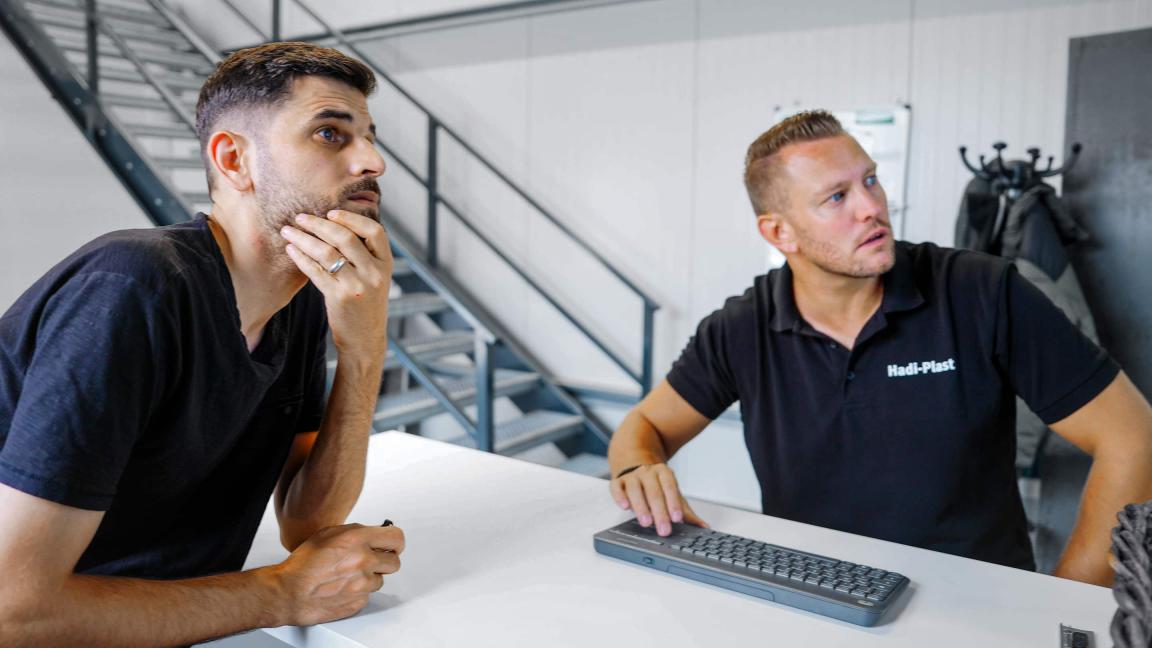 Two men sit at a white desk in a bright room with a metal staircase: on the left, a man with his hand on his chin looks thoughtful, while on the right, a man in a black polo shirt with the "Hadi-Plast" logo works on a keyboard.