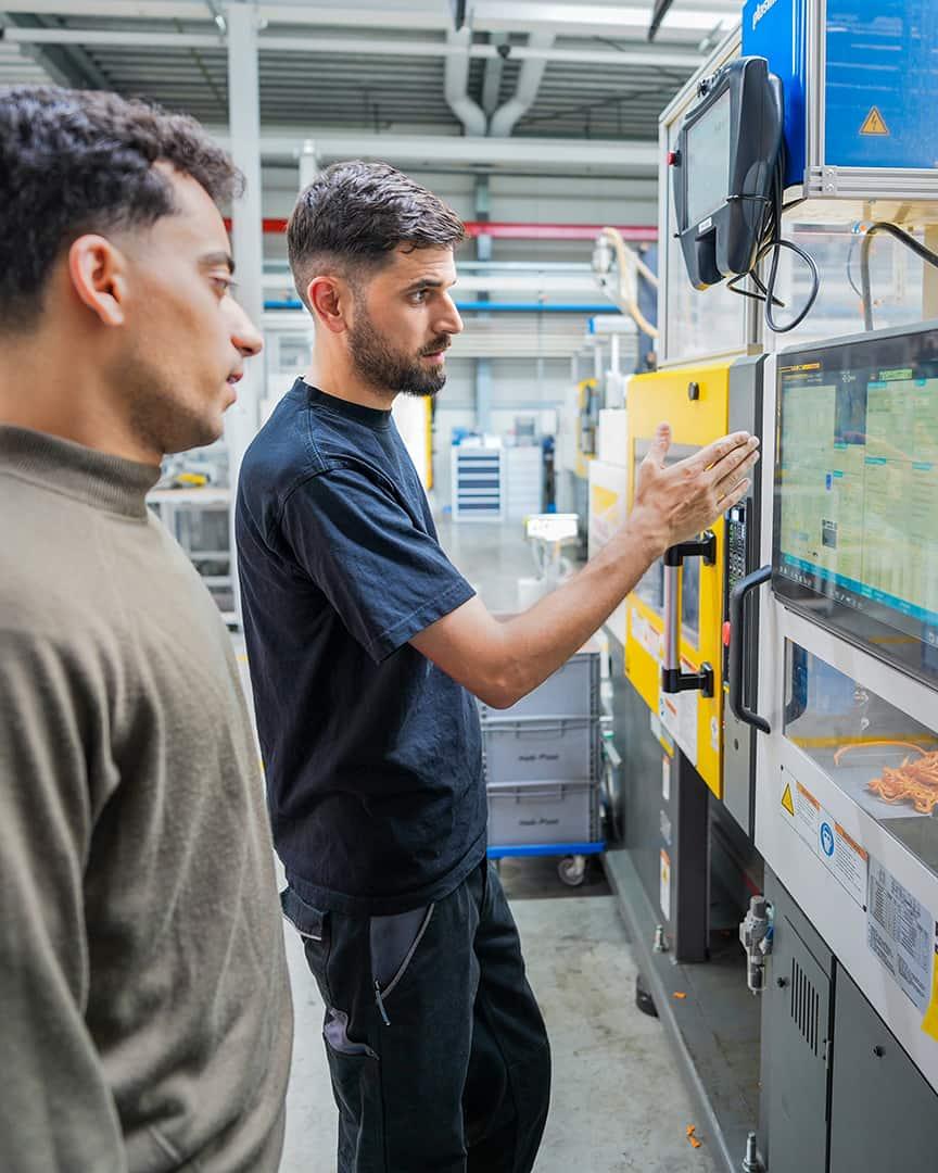 A bearded man in a dark blue T-shirt and black trousers gestures at the control panel of a yellow and white injection molding machine in an industrial hall, while a second man in an olive-green sweater watches attentively.