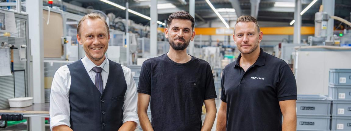 Three men stand side by side in a production hall: on the left, a smiling man in a vest and tie, in the middle, a bearded man in a dark blue T-shirt, and on the right, a man in a black polo shirt with the "Hadi-Plast" logo.