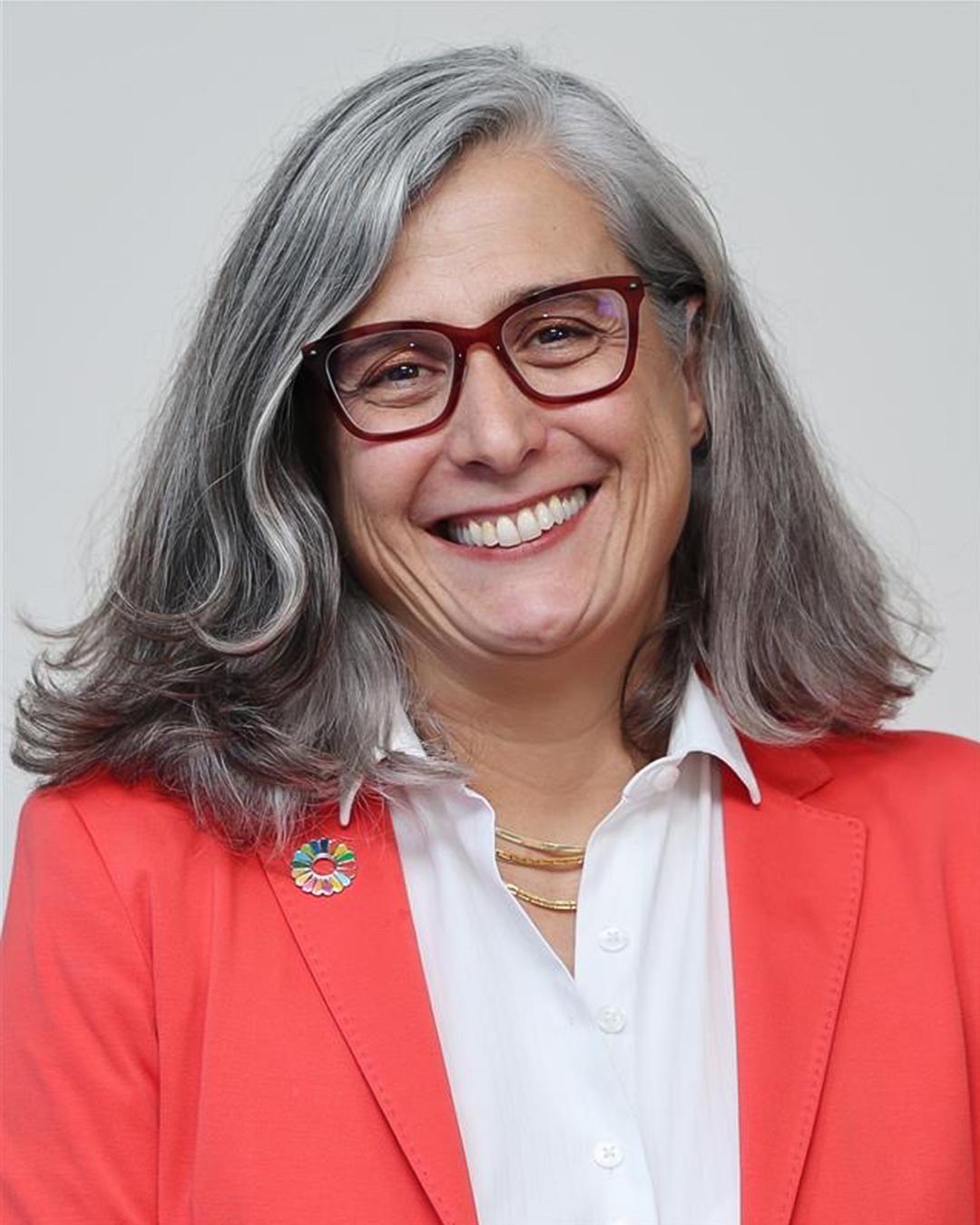 Porträt einer lächelnden Frau mit grauen Haaren und Brille, die ein rotes Sakko über einer weißen Bluse trägt.
