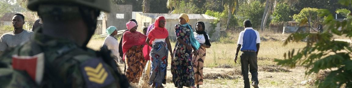 Mehrere Frauen laufen über einen Weg. Ein Soldat steht im Vordergrund und beobachtet die Szene.