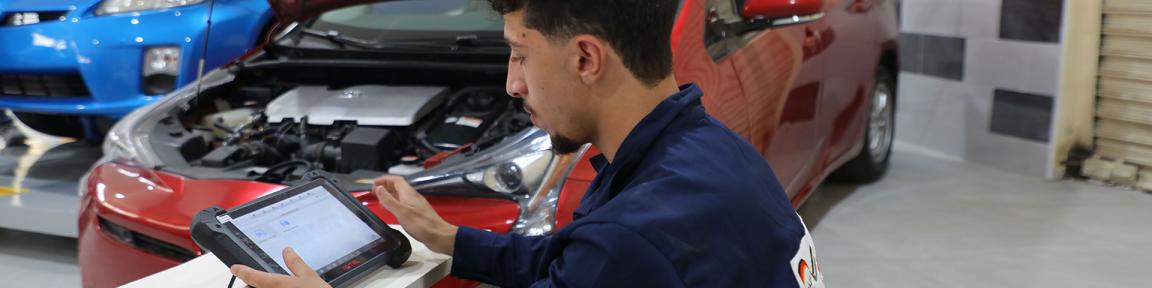 Ein junger Auszubildener prüft in einer Autowerkstatt die Batteriespannung für Hybridfahrzeuge an einem Tablet.