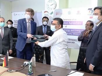 Two men wearing face masks hand over documents to each other. Next to and behind them are six other men; some are clapping. Copyright: GIZ / REEE-SCALE