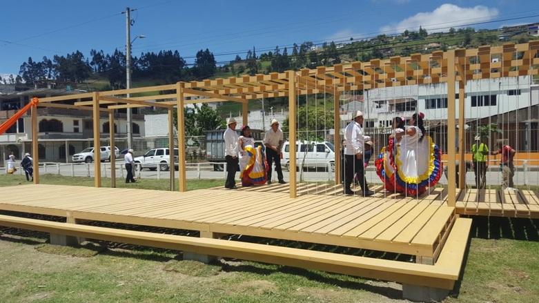 Mujeres vestidas con trajes tradicionales bailan sobre una plataforma de madera en una zona verde de una ciudad.