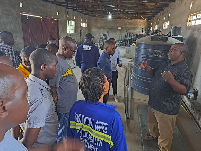 A study group visits a landfill site. They stand in a large room and listen to a speaker.