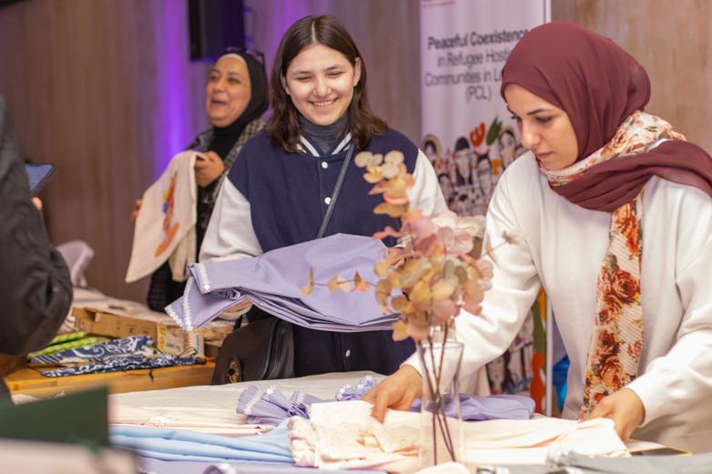 Eine Frau legt an einem Stand genähte Kleider aus. Eine andere Frau reicht ihr weitere Kleidungsstücke an.