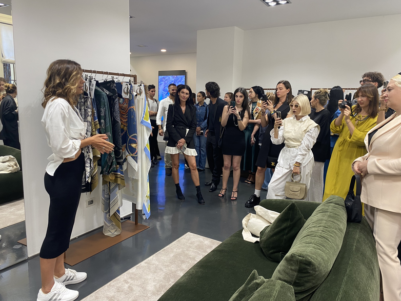 A woman speaks next to a rack of clothes. People stand around her.