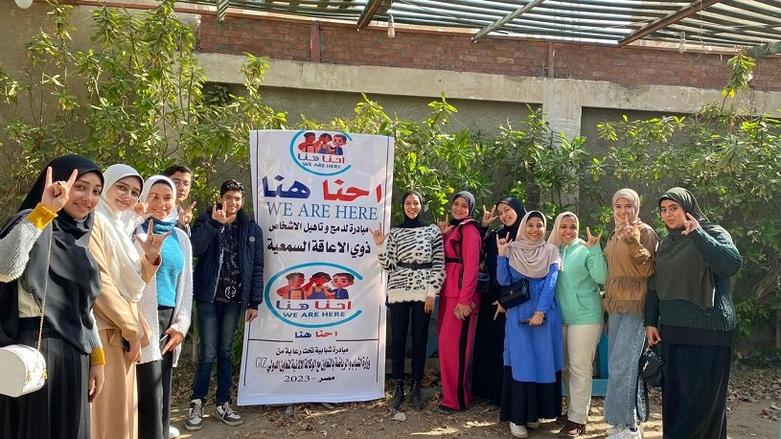 Twelve young people, mainly young women, stand next to a poster and make hand signals, smiling.
