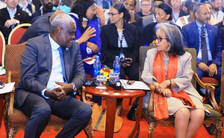 Ein Mann und eine Frau sitzen auf Stühlen vor einem gut besuchten Konferenzsaal und diskutieren.