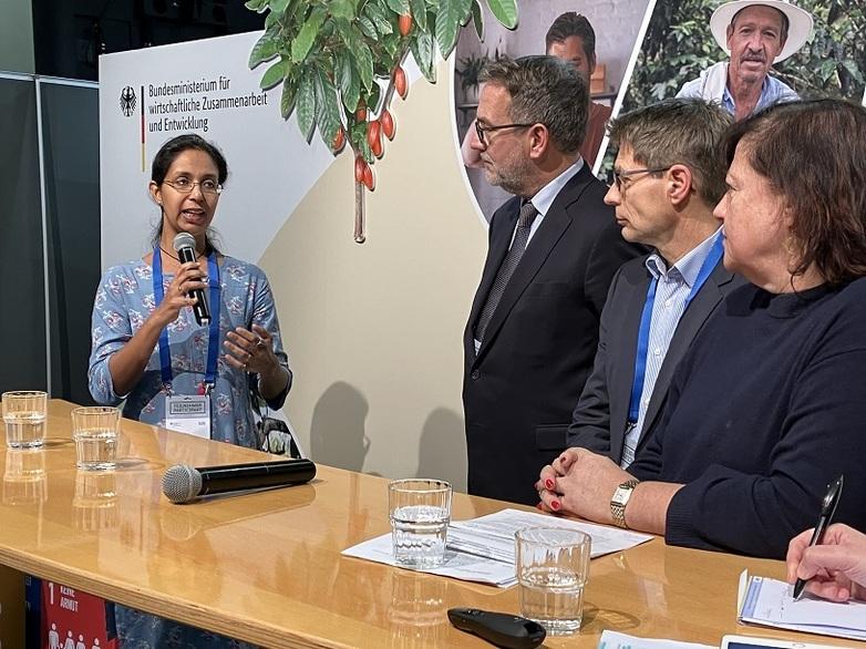A woman with a microphone stands in front of a table and talks to three people.