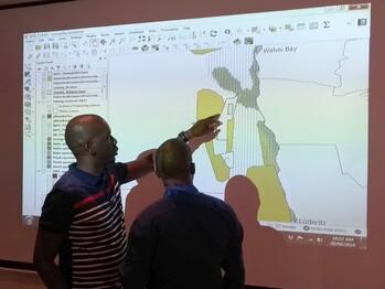 Standing in front of a map projected onto the wall showing the Benguela Current in Namibia, two men discuss biodiversity protection and mining zones. © GIZ MARISMA / Roman Sorgenfrei