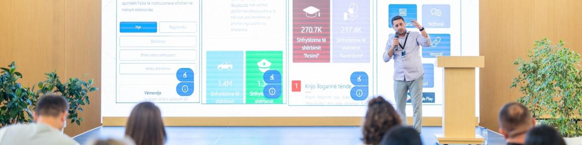 Bei einer Veranstaltung zum Thema E-Governance und Verbesserung der öffentlichen Dienste in Kosovo steht ein Redner auf einer Bühne vor einem großen Bildschirm mit Statistiken zu digitalen Diensten.