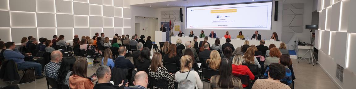 Auf einer Bühne vor einem Publikum sitzen Vertreter*innen. Ein Mann hält eine Präsentation.