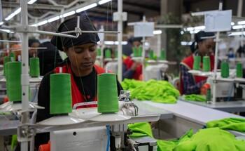 A worker operating a sewing machine surrounded by spools of green thread in a garment factory.