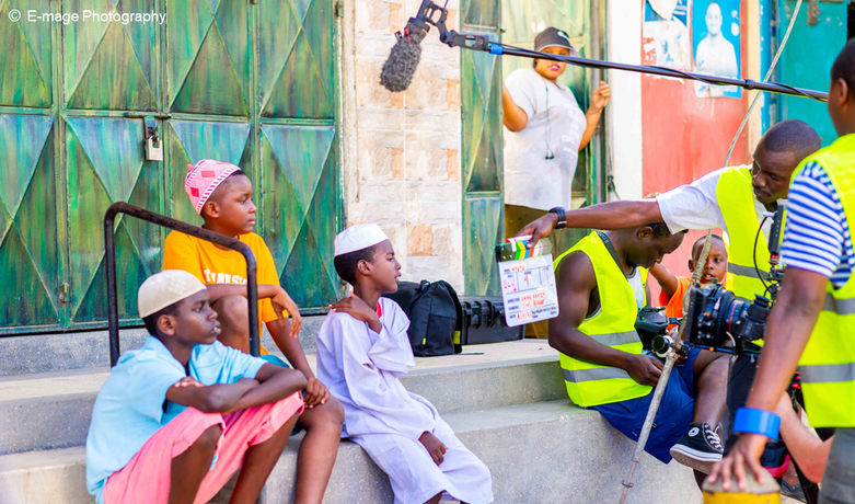A man in a yellow safety vest holding a clapperboard in front of three children while a camera and sound boom are pointed at them.